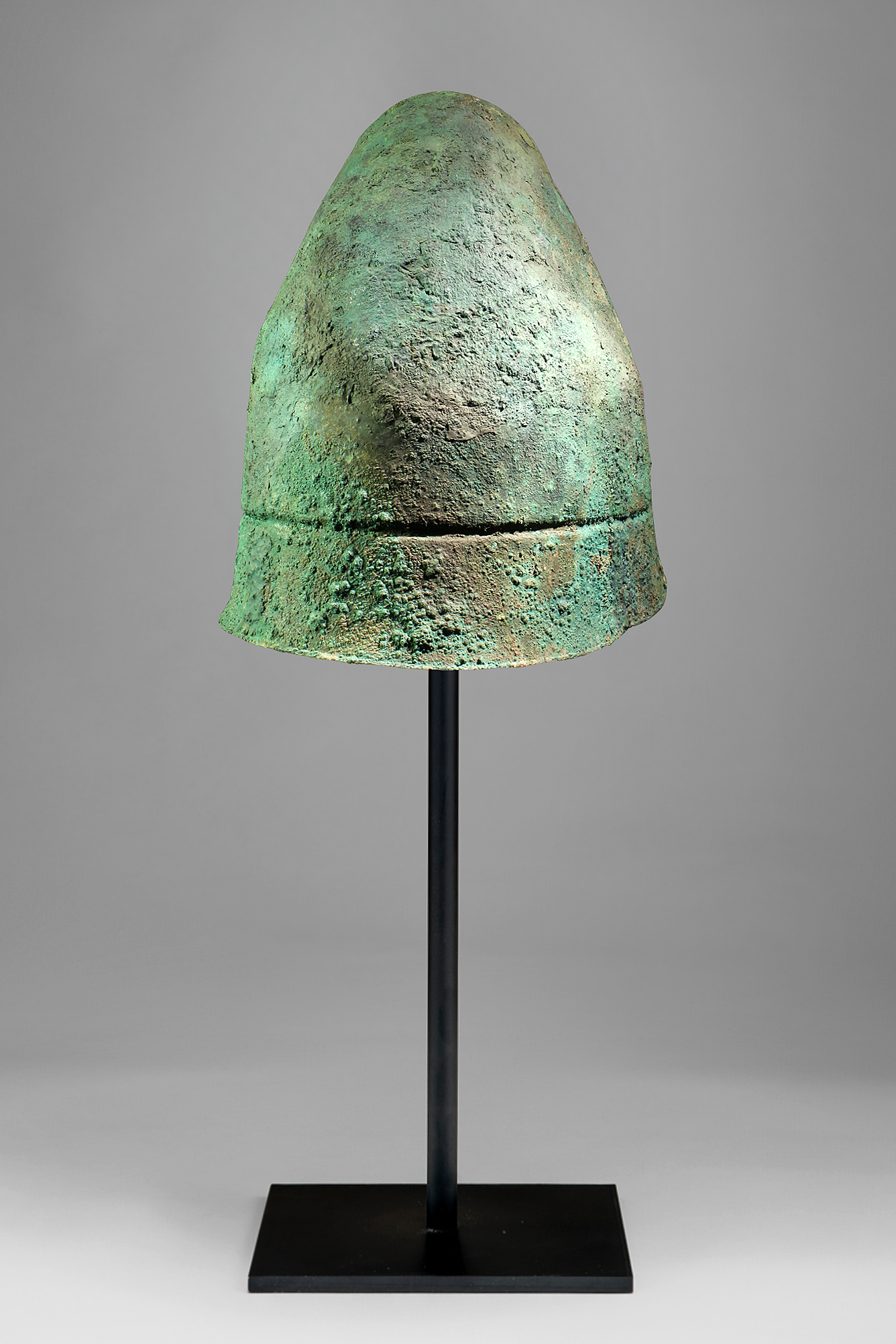 Ancient bronze helmet with a green patina displayed on a stand.