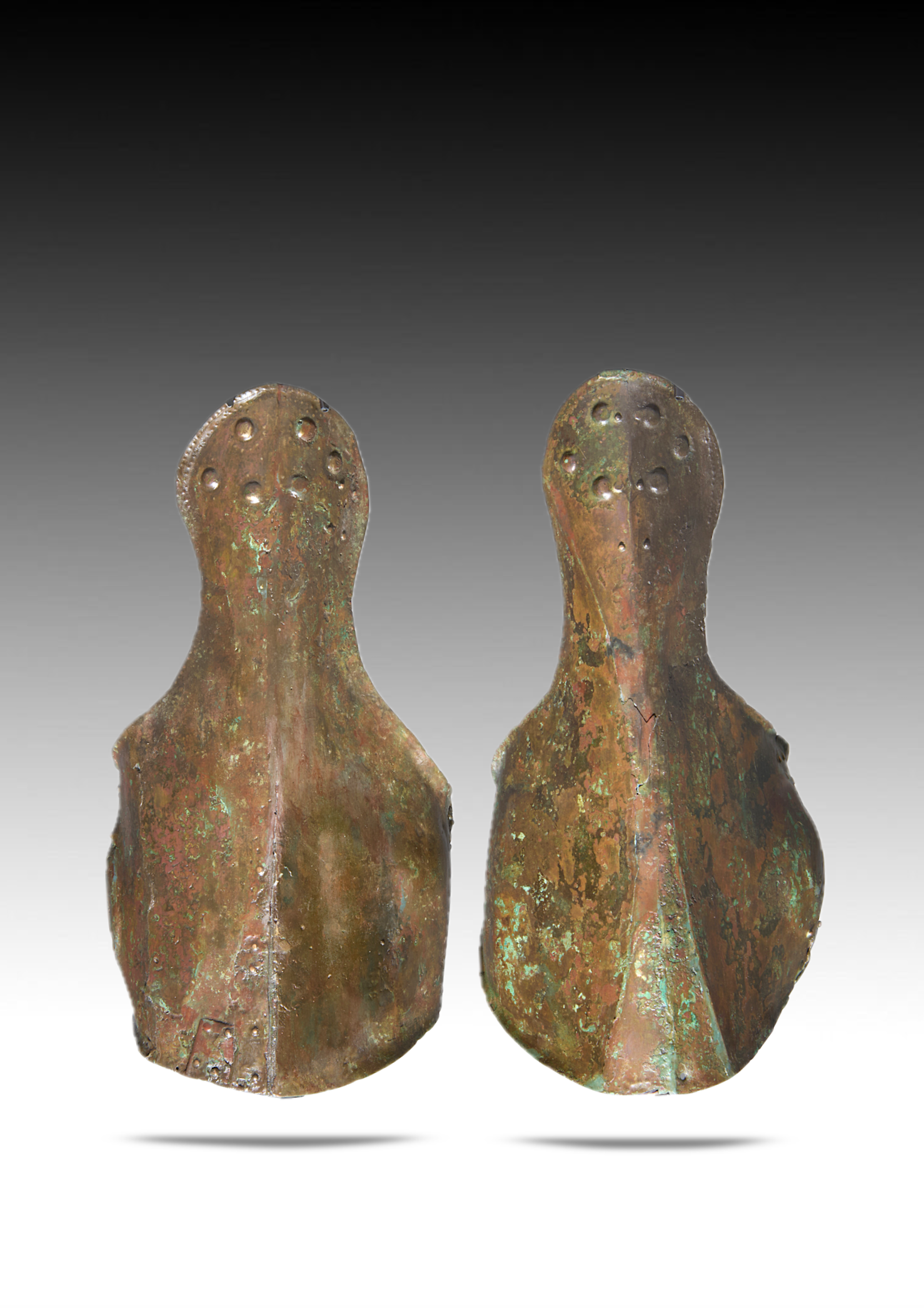 Two weathered, bronze-colored metal figurines with elongated shapes and holes on top, resembling stylized human forms, standing on a white surface with shadows below.
