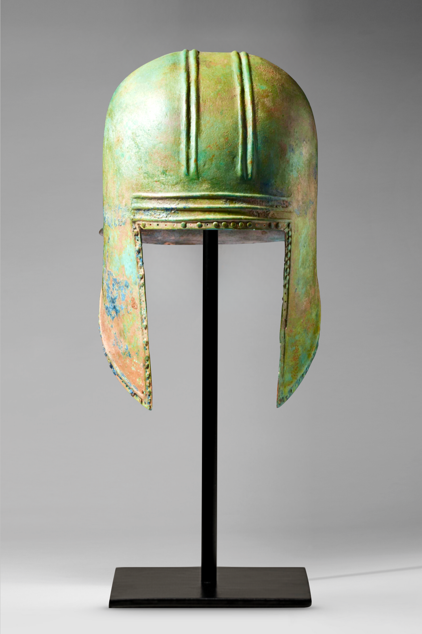 A bronze helmet with a green patina, displayed on a black stand. The helmet has a rounded top and elongated side extensions, with rivets along the edges.