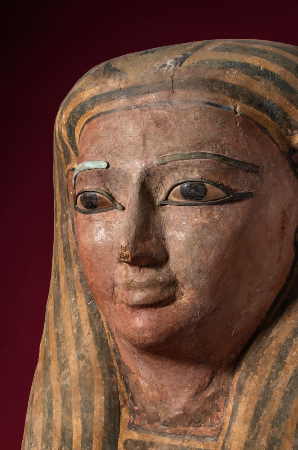 Close-up of an ancient Egyptian painted wooden sculpture of a woman with a serene expression, wearing a striped headdress, against a dark red background.