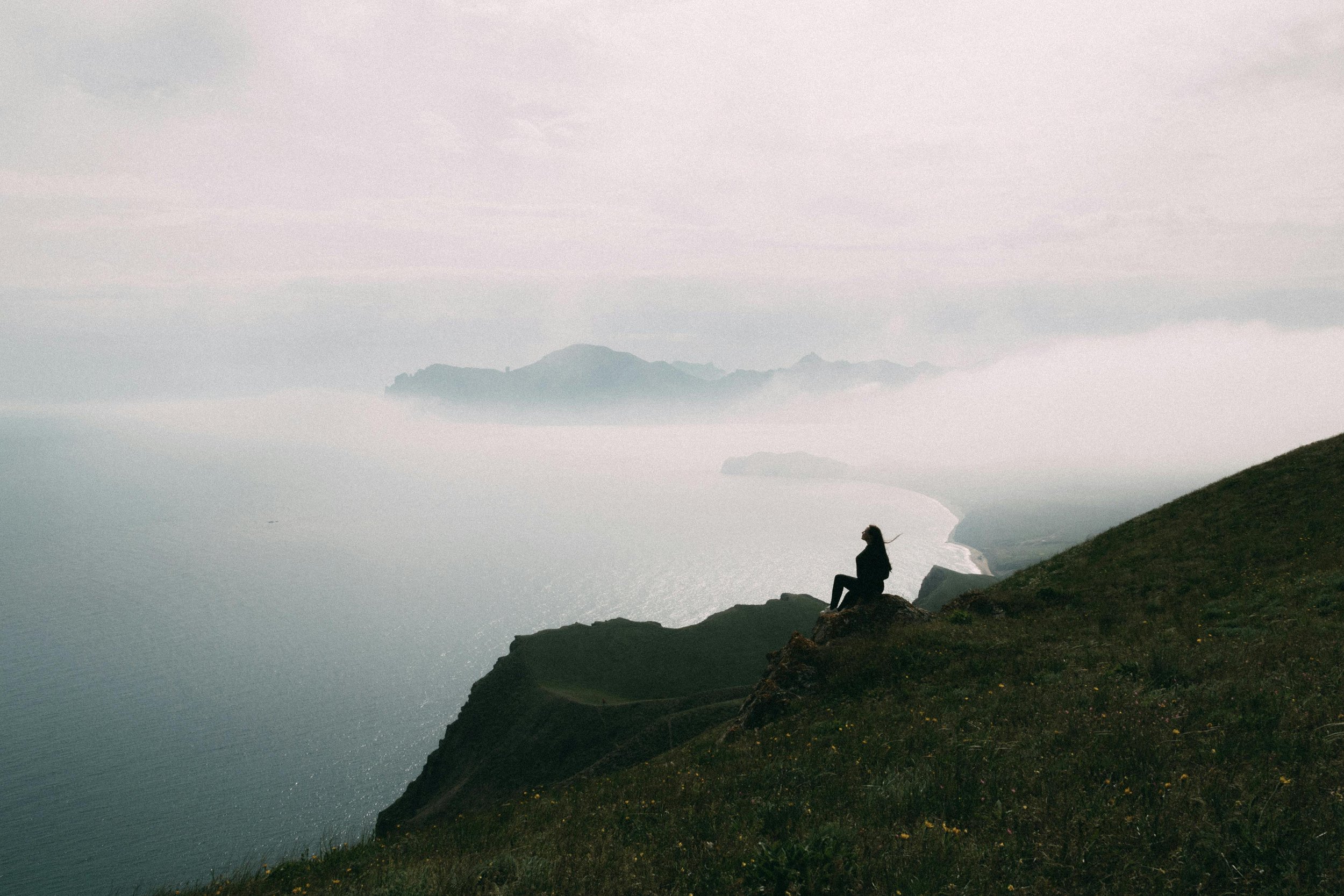 Person sitting on a grassy hillside overlooking the ocean, with misty mountains in the background