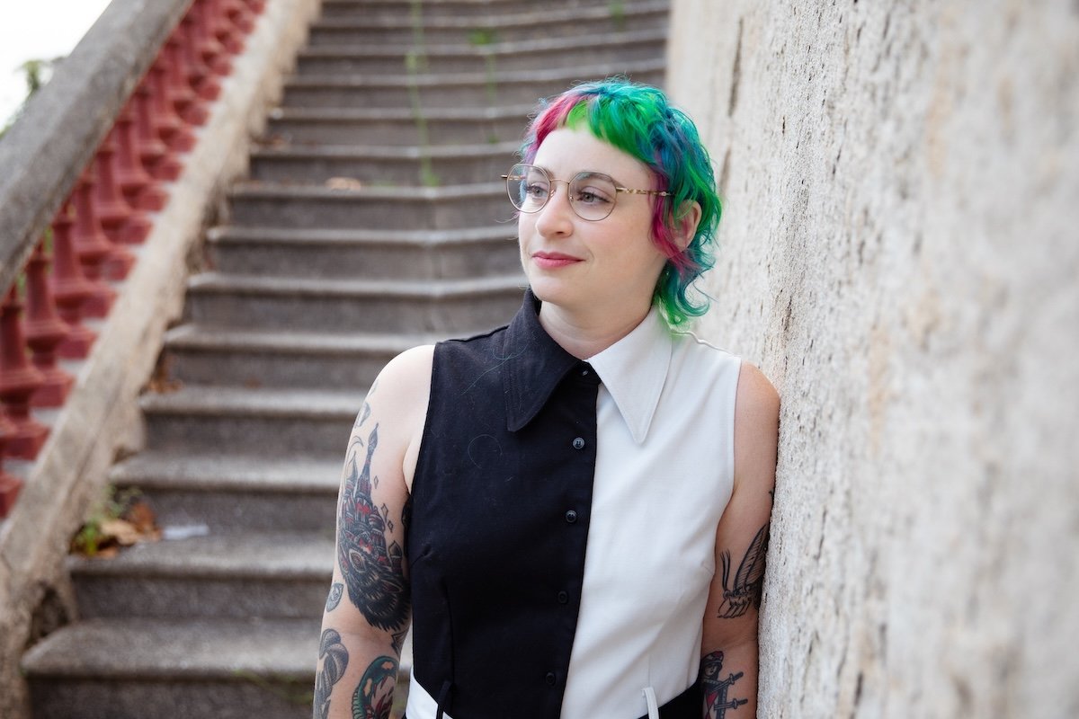 A woman with colorful hair, wearing glasses, leaning against a wall near outdoor stairs.