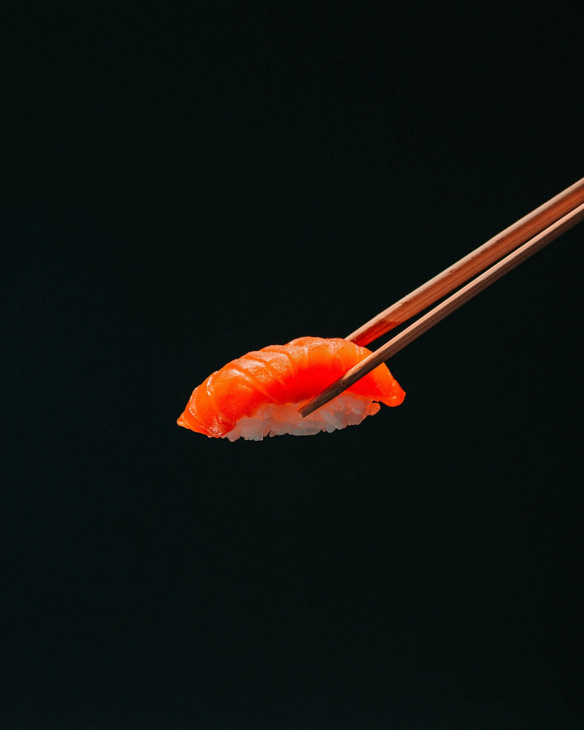 A piece of salmon nigiri sushi held with wooden chopsticks against a dark background.