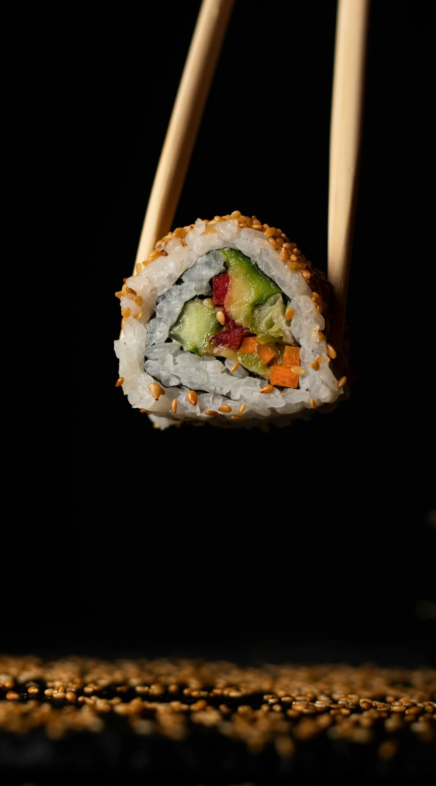 Close-up of a sushi roll held by chopsticks, with rice, avocado, cucumber, and carrots inside, topped with sesame seeds, against a black background.