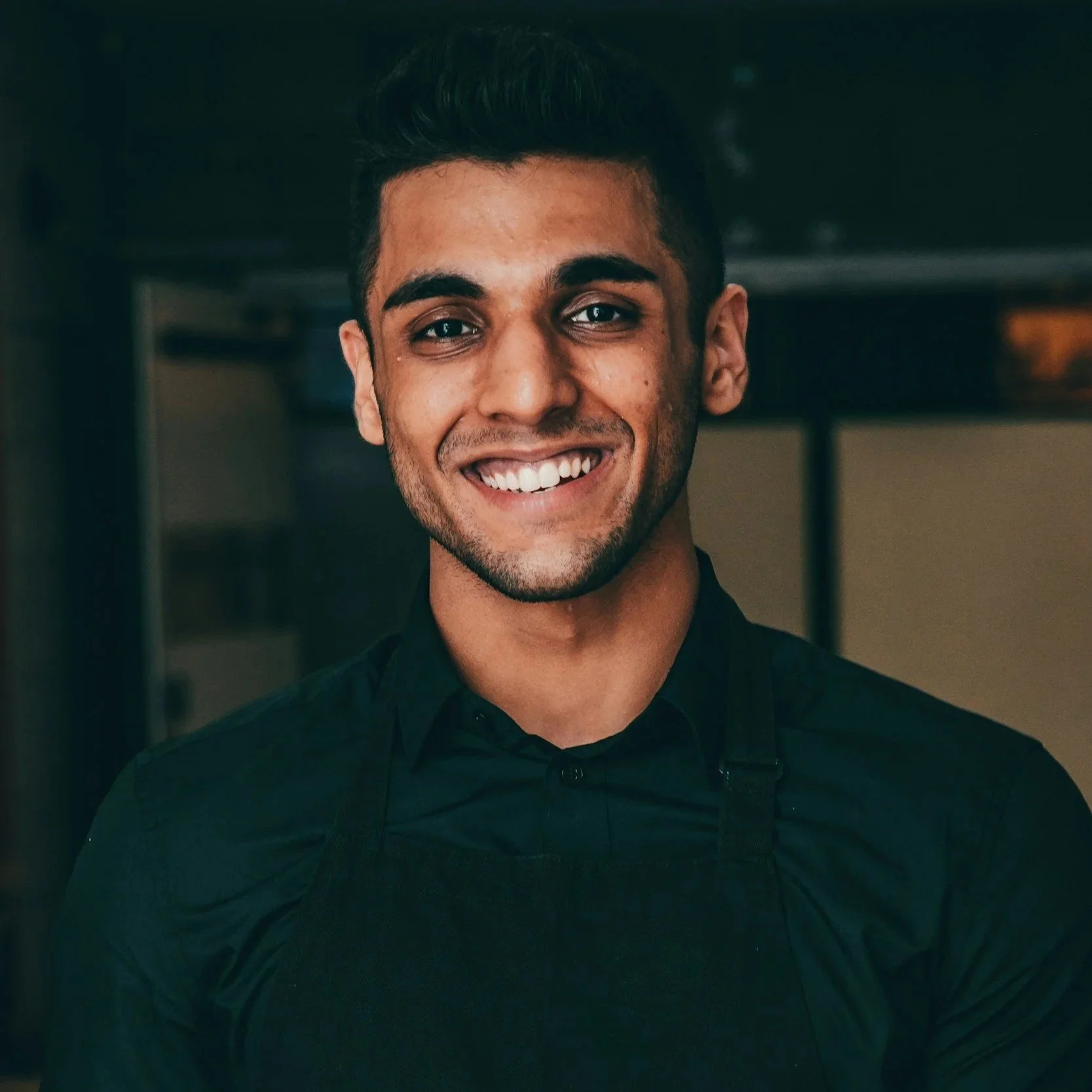 Young man with dark hair smiling in a black shirt, likely working in a kitchen or restaurant.