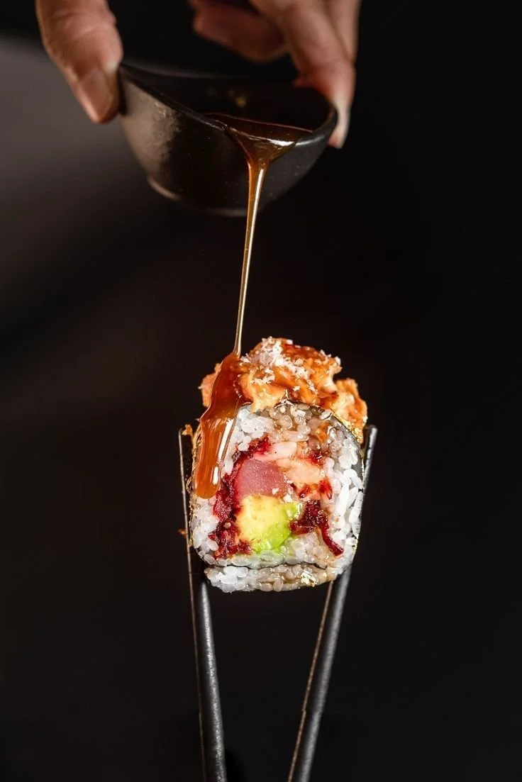 Sushi roll with white rice, avocado, and raw fish, topped with sauce being drizzled from a small dark bowl, against a black background.