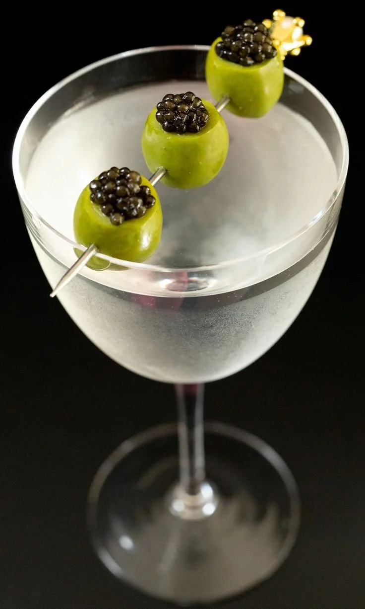 A cocktail in a tall, elegant glass garnished with three green olives threaded onto a skewer, each topped with black caviar, and a small gold crown decoration on the last olive.