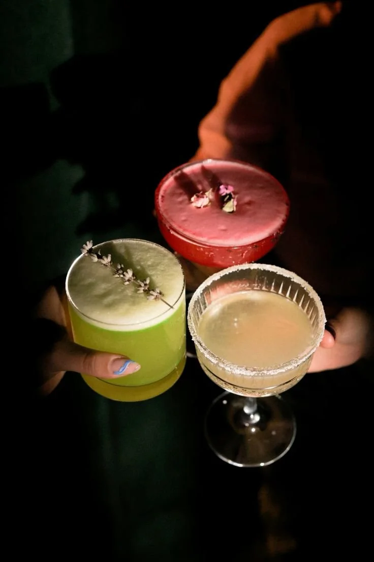 Three cocktails being held up in a dark setting. One green with a flower garnish, one pink with pieces of fruit, and one pale yellow in a coupe glass with a salted rim.