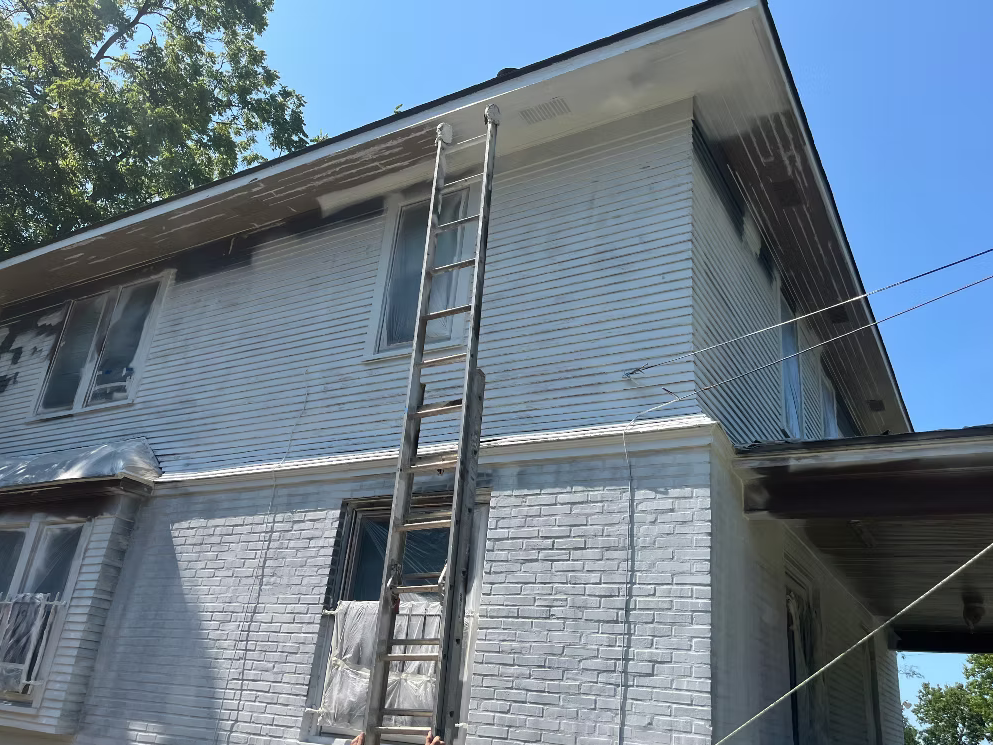 Ladder leaning against the side of a house with white brick and vinyl siding, with some wet paint on the trim near the roof, under a clear blue sky.