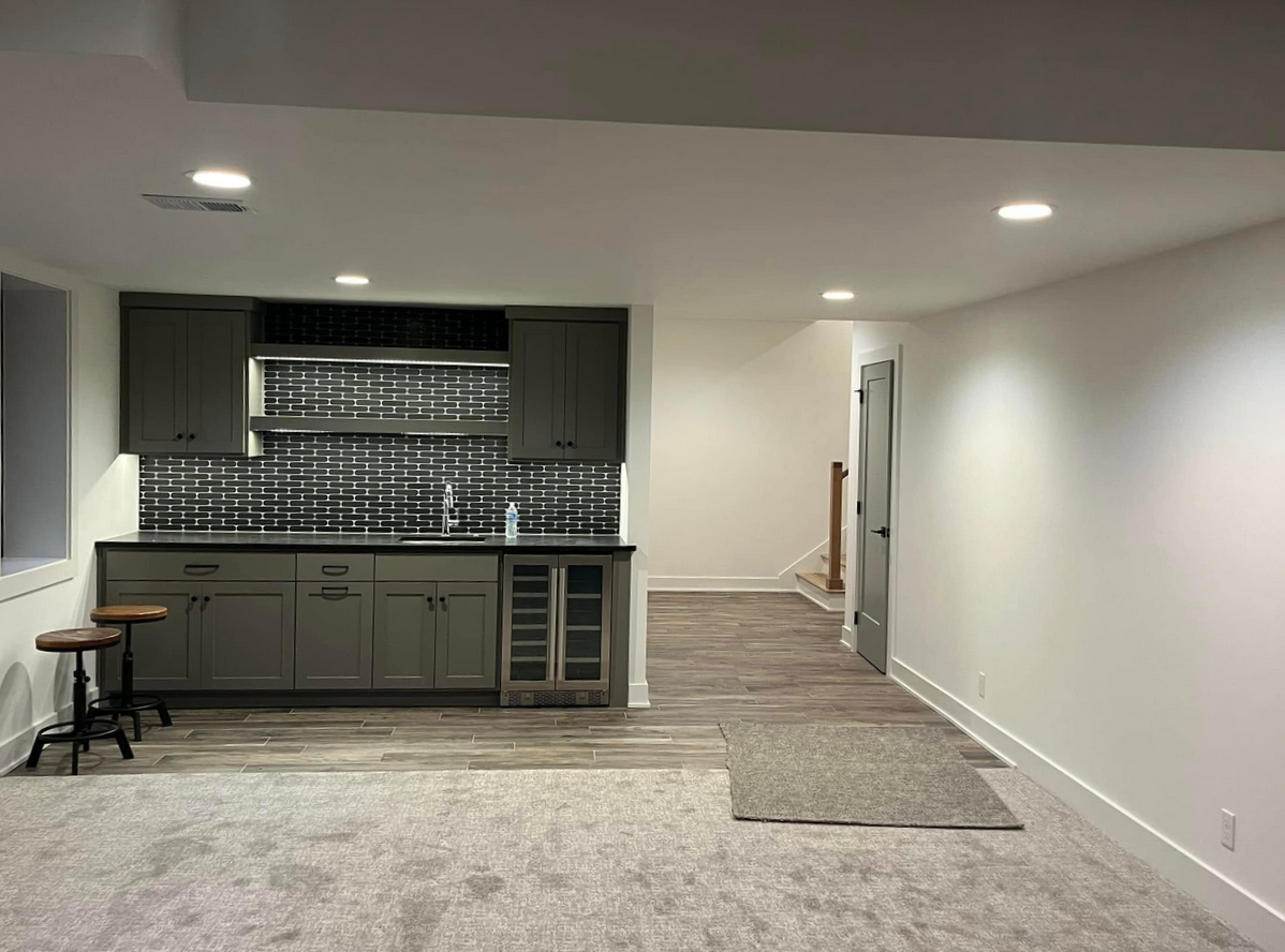 Empty basement room with kitchenette featuring gray cabinets, black subway tile backsplash, and small fridge with glass door, illuminated ceiling lights, and no furniture or decorations.
