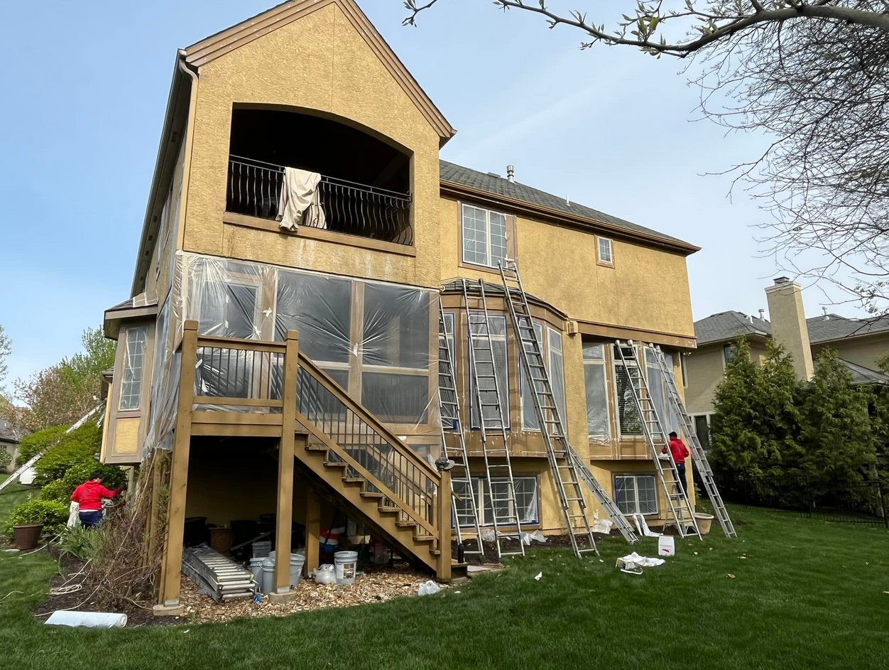The back of a large two-story house under renovation with workers installing or repairing siding. Several ladders lean against the house, and work materials are visible underneath the staircase and around the yard.