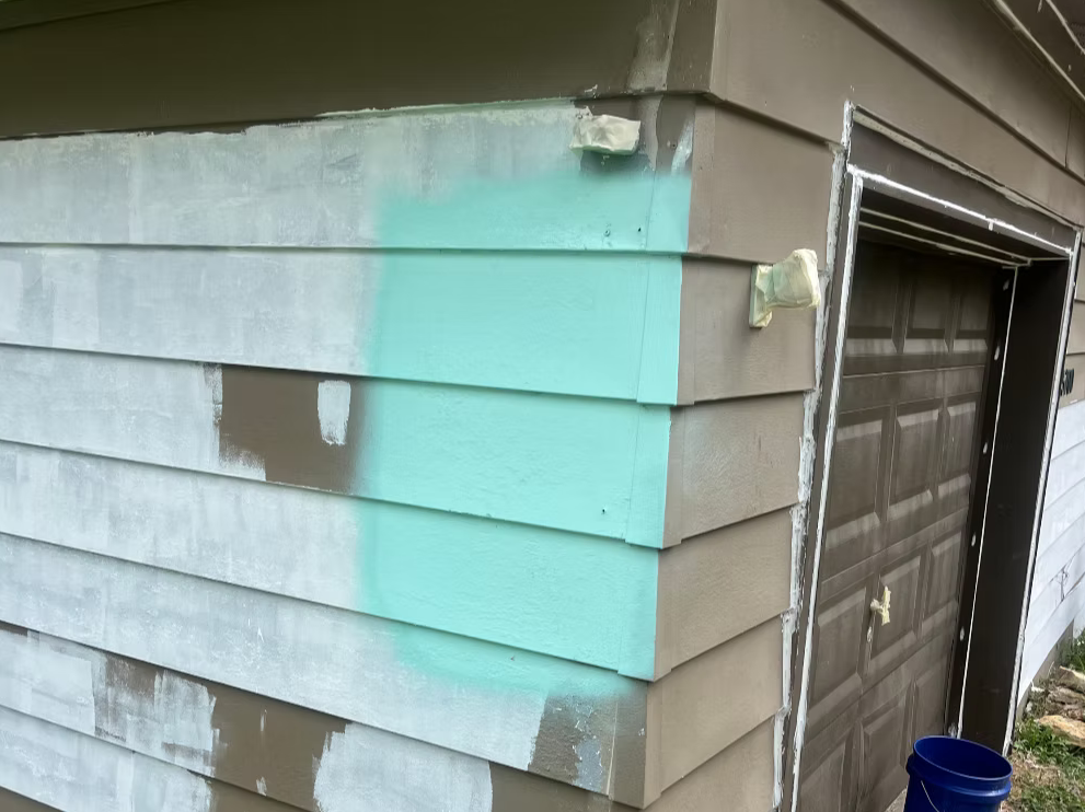 Corner of a house with beige horizontal siding, with some sections painted in light turquoise and others covered in primer. A blue bucket is on the ground near the house.