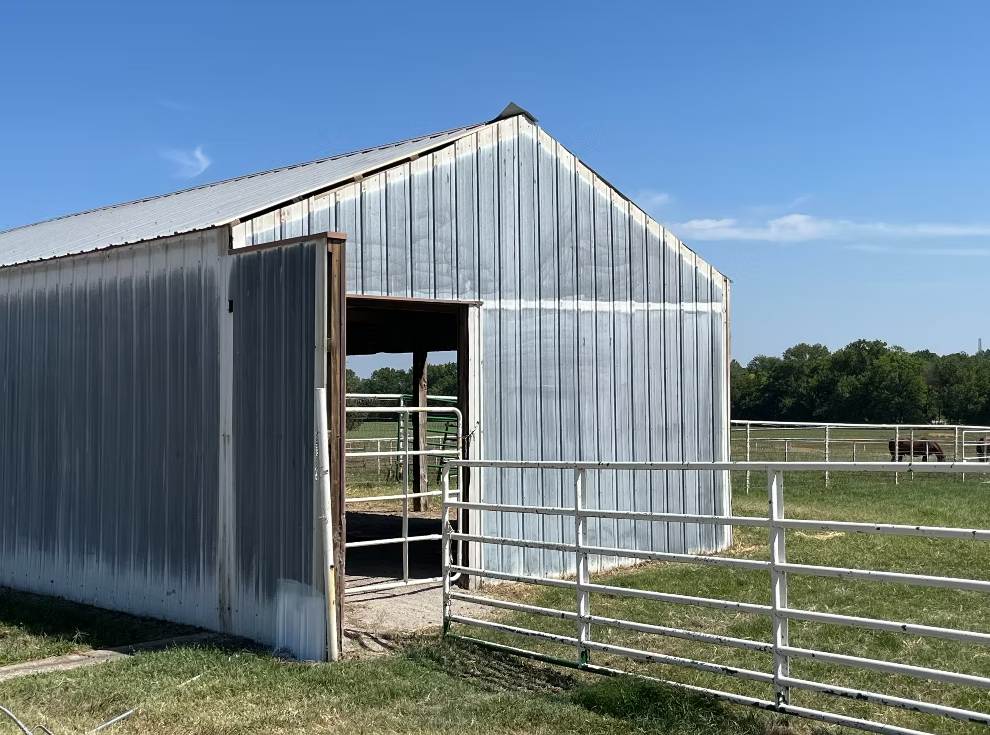 A metal barn with an open door on a rural farm, surrounded by fenced pastures and a clear blue sky.