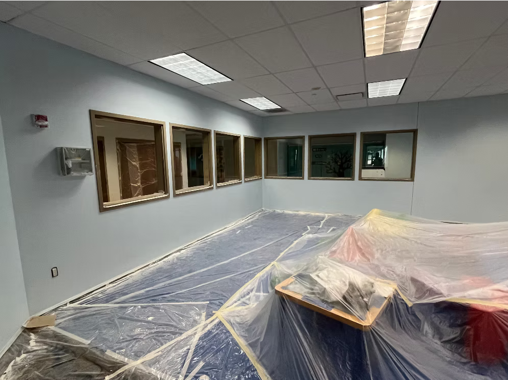 Empty room with construction plastic covering the floor and furniture, and seven windows on the wall.