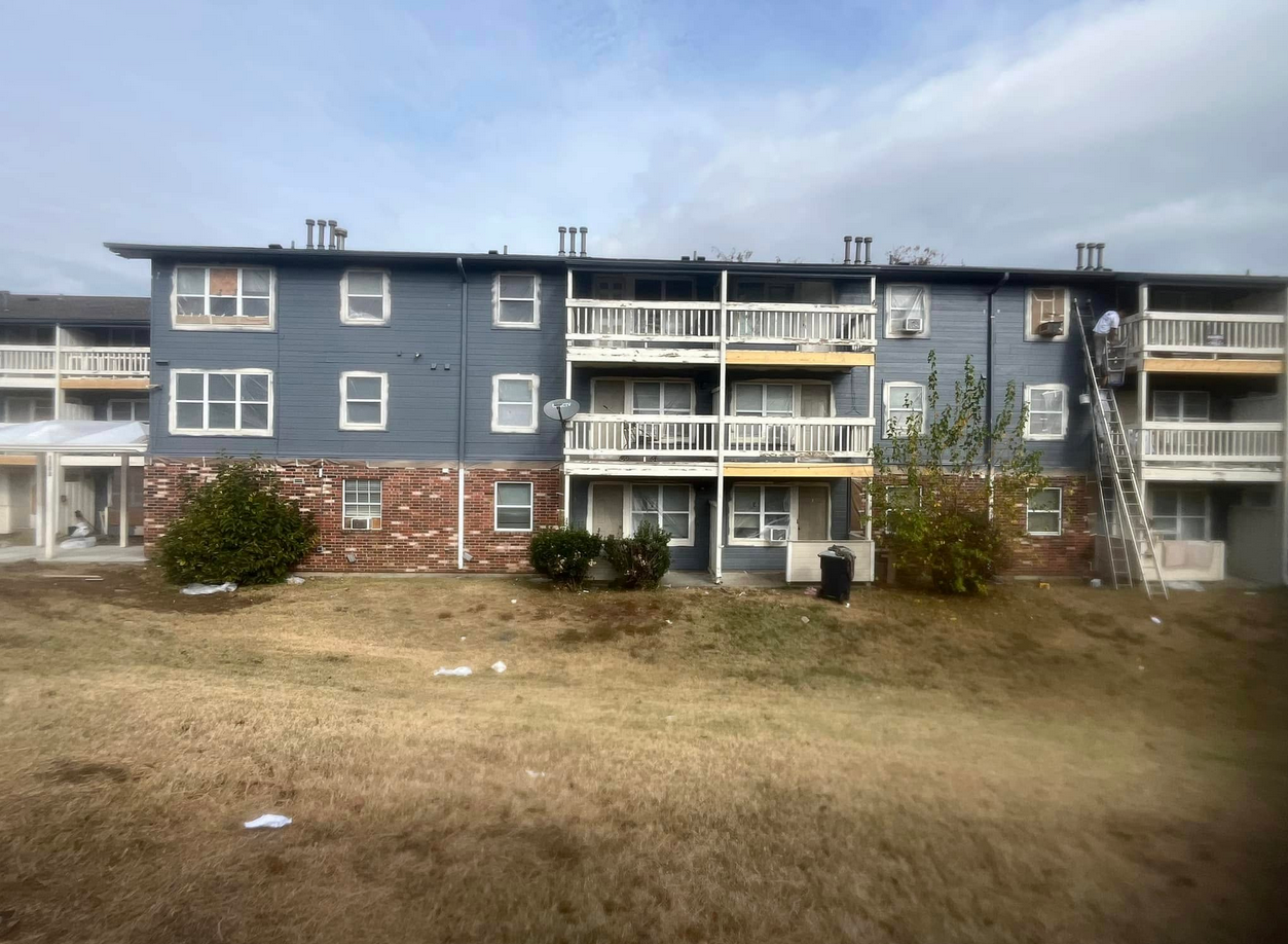 Multi-story residential building with blue siding, brick foundation, and balconies, with a person on a ladder cleaning upstairs.
