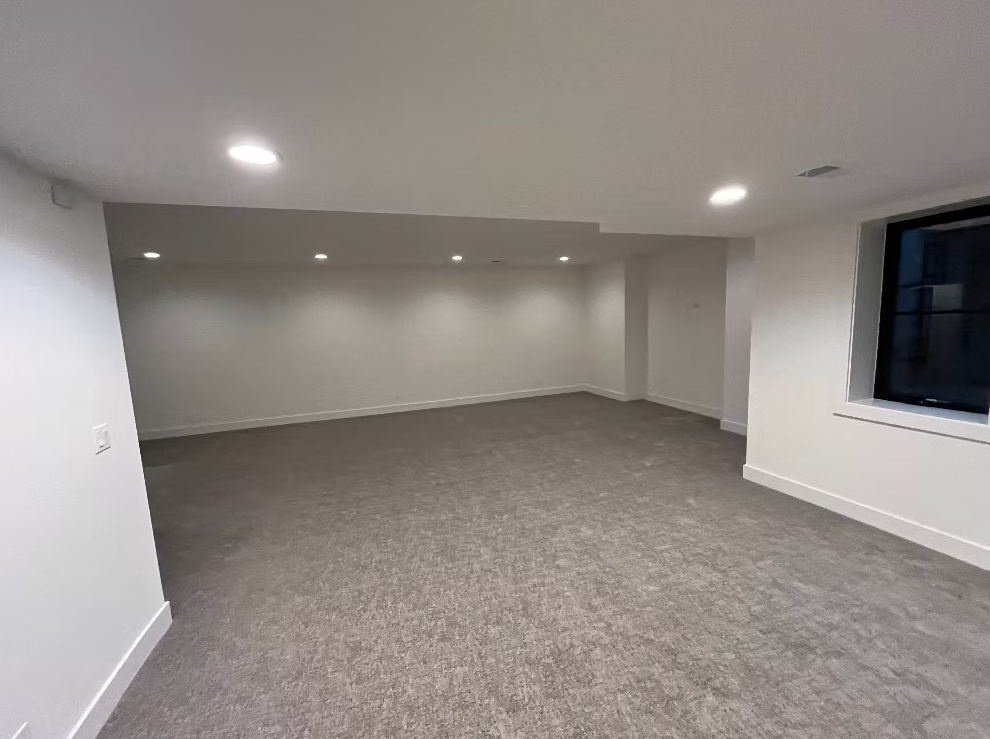 Empty basement room with white walls, gray carpet, recessed ceiling lights, and a window.