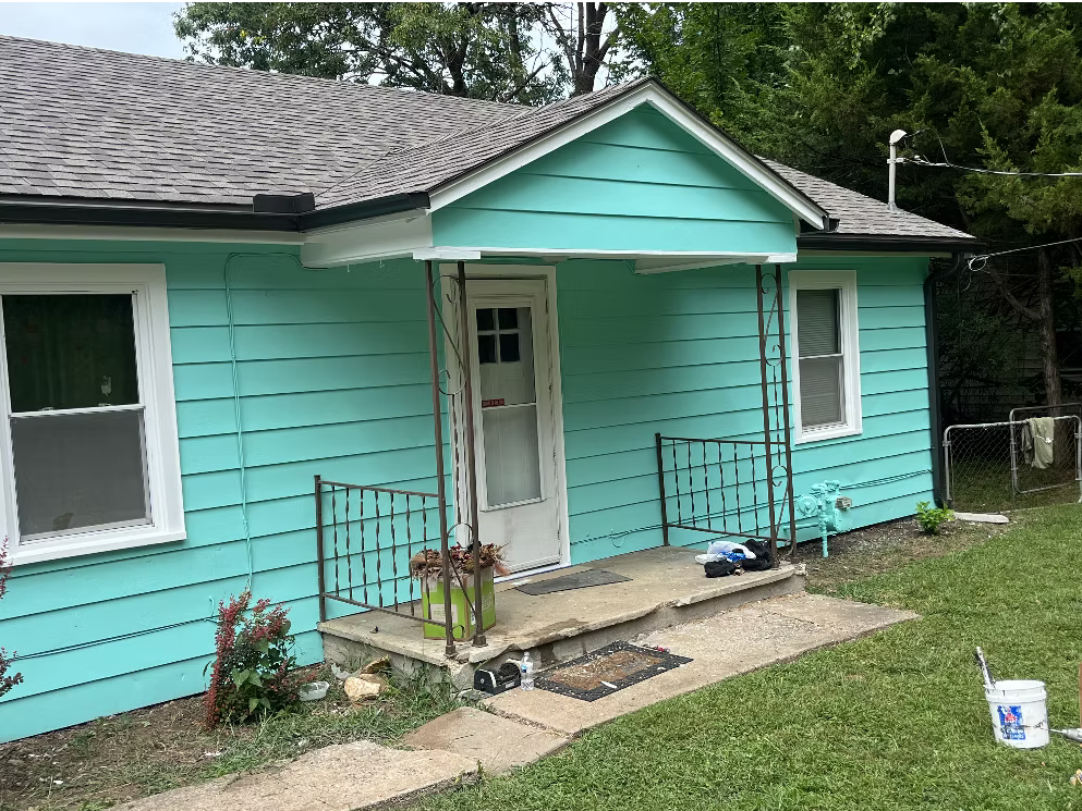 A small turquoise house with white trim and a small front porch with two metal railings. There is a concrete walkway leading to the porch and a yard with grass and some plants. A white house door and two windows are visible, and there are some outdoor items near the porch, including a potted plant, a bucket, and miscellaneous objects. Trees are in the background.