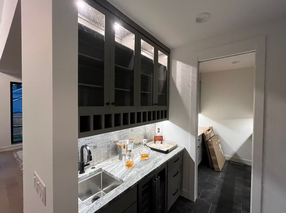 A small bar area with a granite countertop, glass-front upper cabinets, and a sink. There is a bottle of liquor and glasses on the counter, and a doorway leading to another room with stacked boxes.