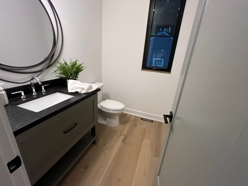 A small bathroom with a white toilet, a gray vanity with a black countertop, a clear glass soap dispenser, a white towel, a green potted plant, and a rectangular mirror on the wall. There is a window showing a house exterior, and the floor is light-c