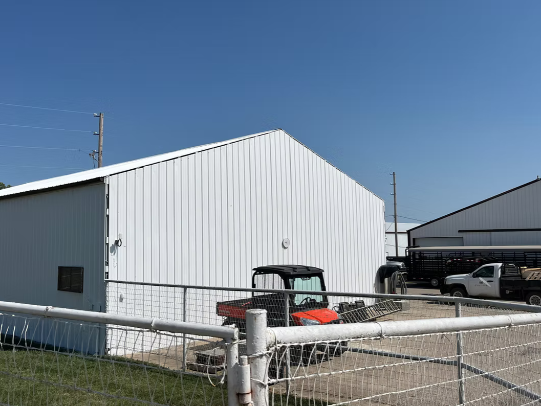 White agricultural or industrial building with a gravel lot, a red utility vehicle, and several trucks parked nearby, surrounded by a chain-link fence under a clear blue sky.