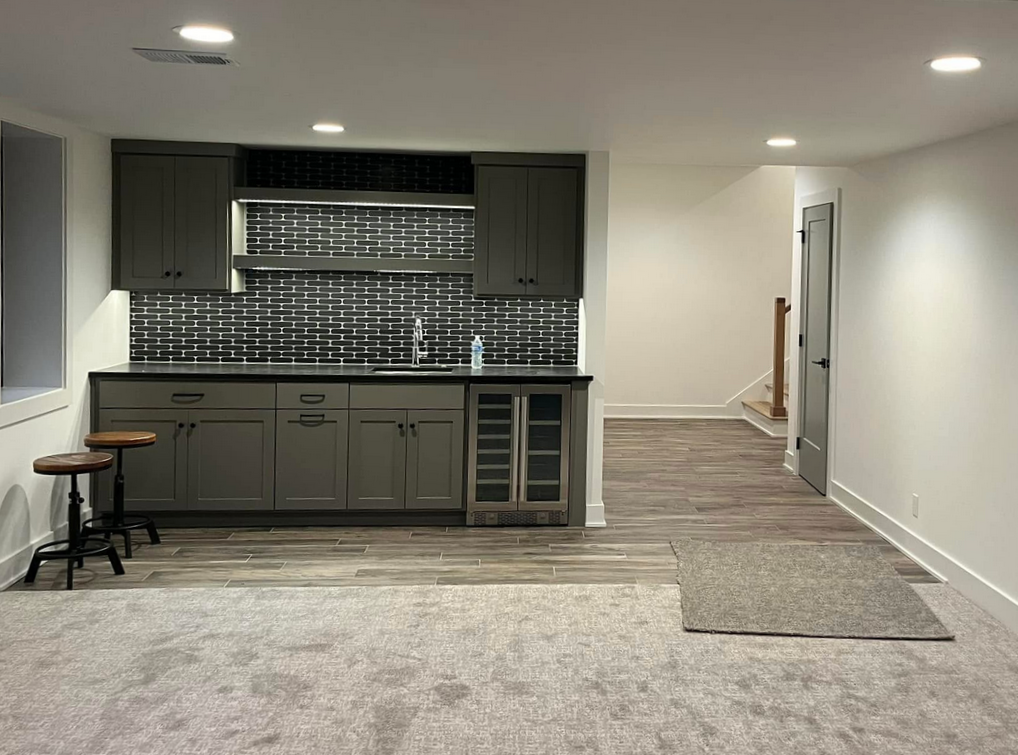 A basement room with a kitchenette featuring grey cabinets, black subway tile backsplash, a small sink, and a wine fridge. There are two stools on the left, wood-look flooring, and a beige area rug.