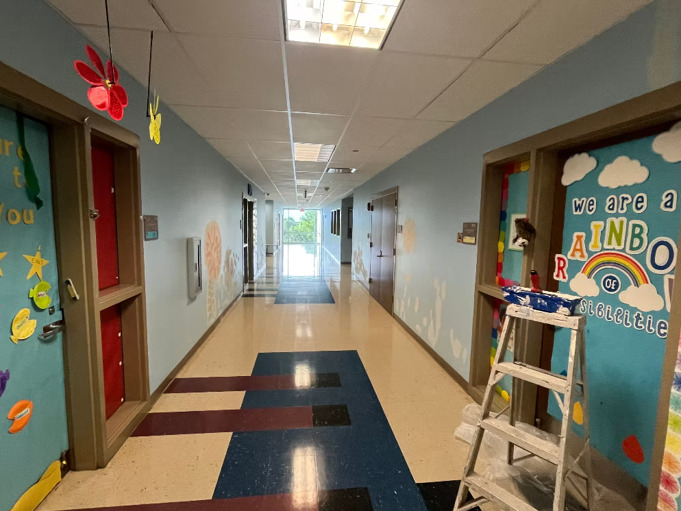 Colorful hallway with decorations and artwork, including a rainbow and clouds, with gates and a ladder in the foreground and a door on the right side.