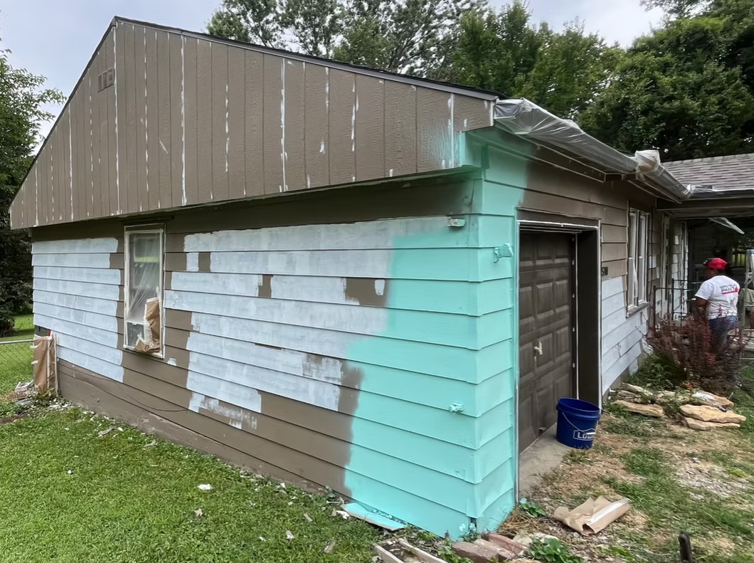House exterior with ongoing siding paint renovation, partially painted in light bluish-green, others in light brown, workers present.