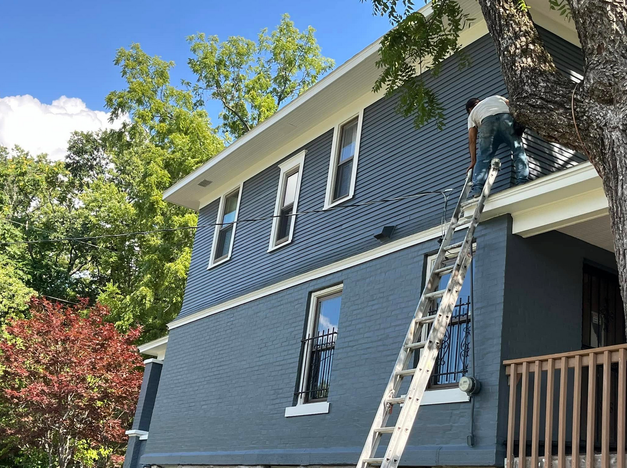 A man standing on a ladder painting the upper part of a house exterior with blue siding. The house has white trim around the windows, a small porch with a wooden railing, and is surrounded by trees with green and red leaves under a blue sky.