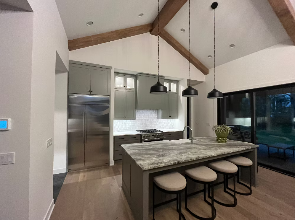 Kitchen with gray cabinets, stainless steel refrigerator, white subway tile backsplash, stovetop, granite island with sink, four barstools, pendant lights, sliding glass door, and a plant on the island.