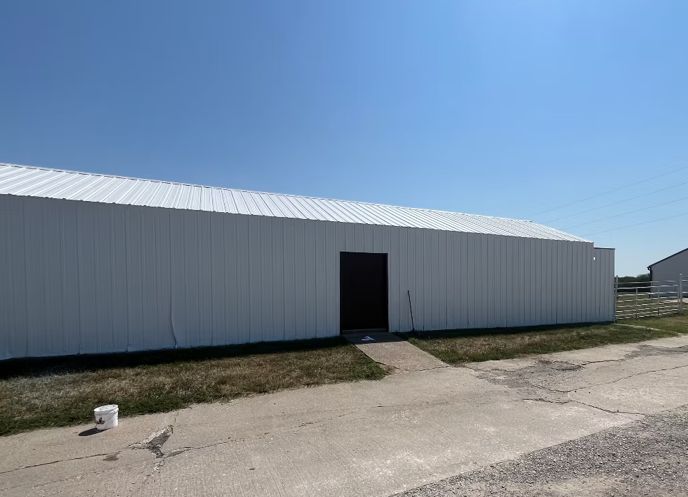 A long white metal building with a sloped roof, a small doorway in the middle, and a concrete walkway leading to the entrance, situated on a partly grassy area with a clear blue sky overhead.
