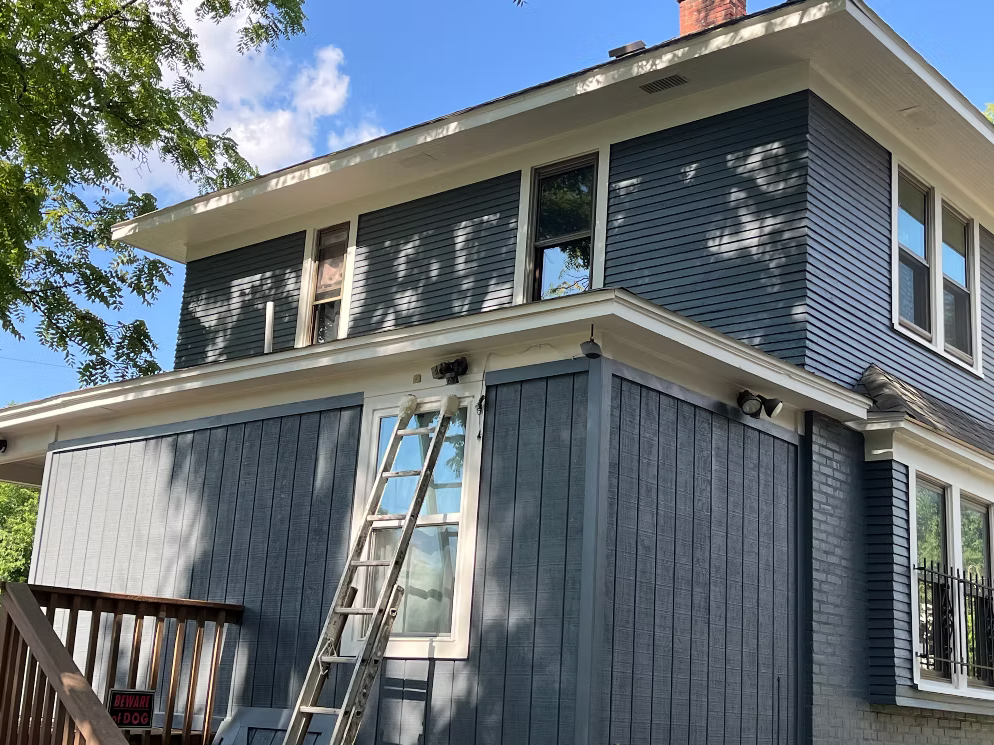 A two-story house with blue siding, white trim, multiple windows, and a ladder leaning against the front wall. There are trees and a partly cloudy sky in the background.