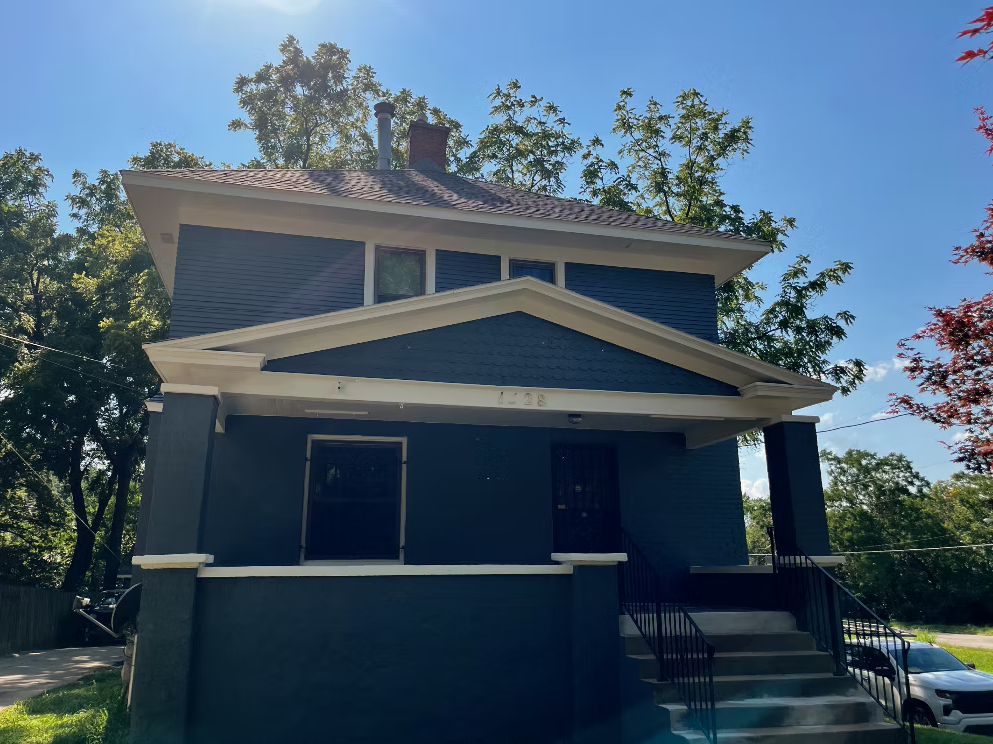 Two-story blue house with a front porch, steps, and black railing, surrounded by trees and a blue sky.