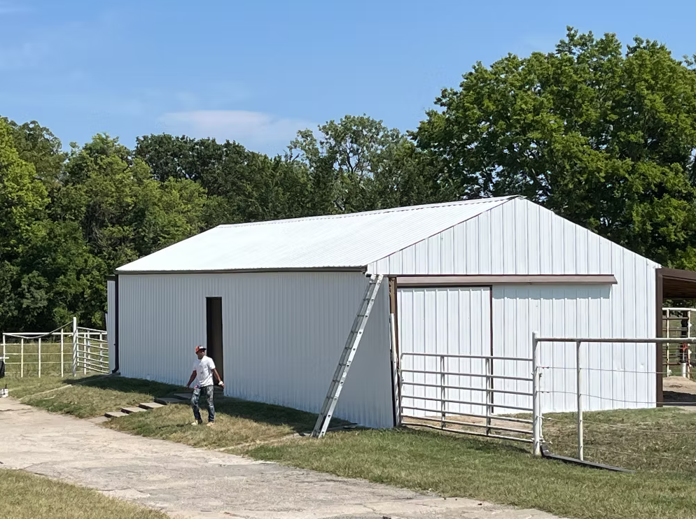 A white metal building on a grassy area with trees in the background, a man wearing a hat standing nearby, a ladder leaning against the building, and a partially open sliding door.