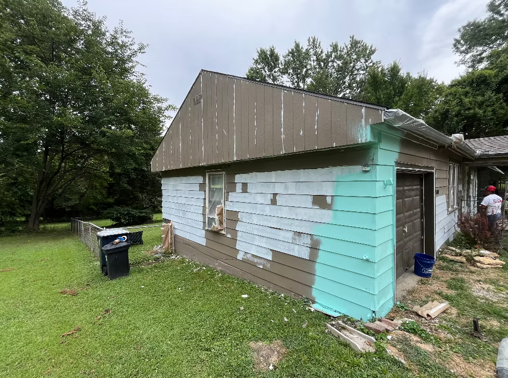 House with siding being painted light blue, with painters working on the exterior, in a green yard with trees and a trash bin nearby.