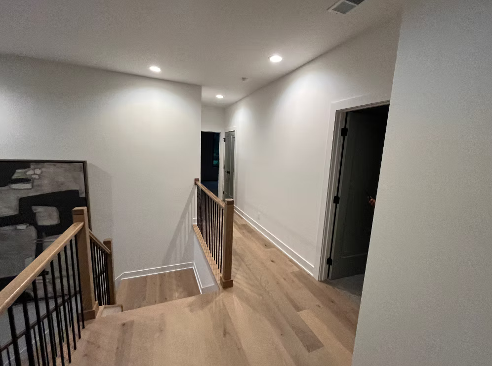 A staircase with wooden handrails leading down to a hallway in a modern home with white walls, hardwood floors, and recessed lighting.