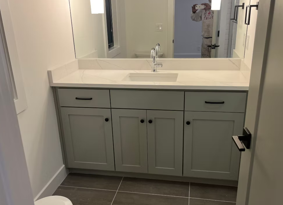 Bathroom vanity with a large mirror, white countertop, and gray cabinets, with a person taking a mirror selfie.