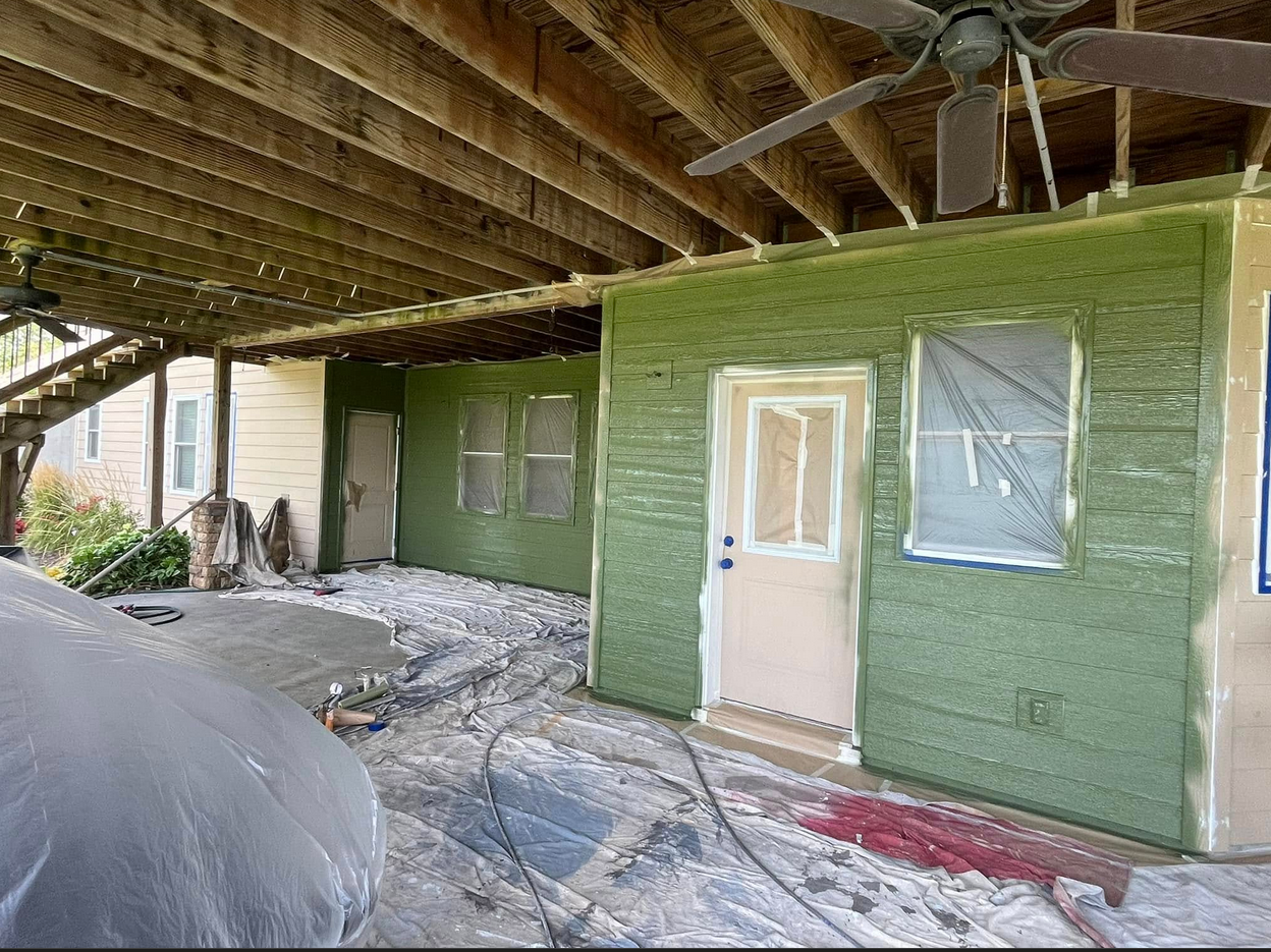 Underneath a wooden deck, a small building is being painted green, with plastic covering the floor and windows to protect them during painting. Construction materials and tools are scattered on the ground.