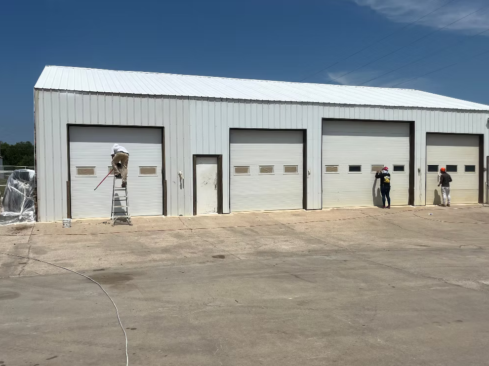 Four workers painting or cleaning the garage doors of a large metal storage building on a clear day.