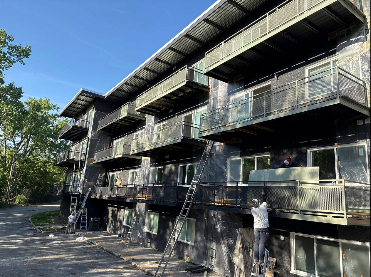 Construction workers on ladders install or repair building panels on a multi-story apartment complex with black exterior walls and metal balconies, surrounded by trees on a sunny day.