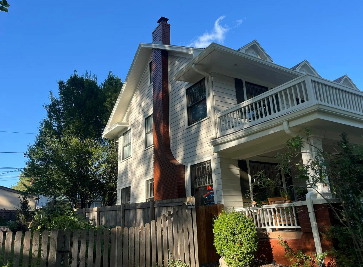 A multi-story white house with a red brick chimney and a balcony, surrounded by green trees and a wooden fence, under a clear blue sky.