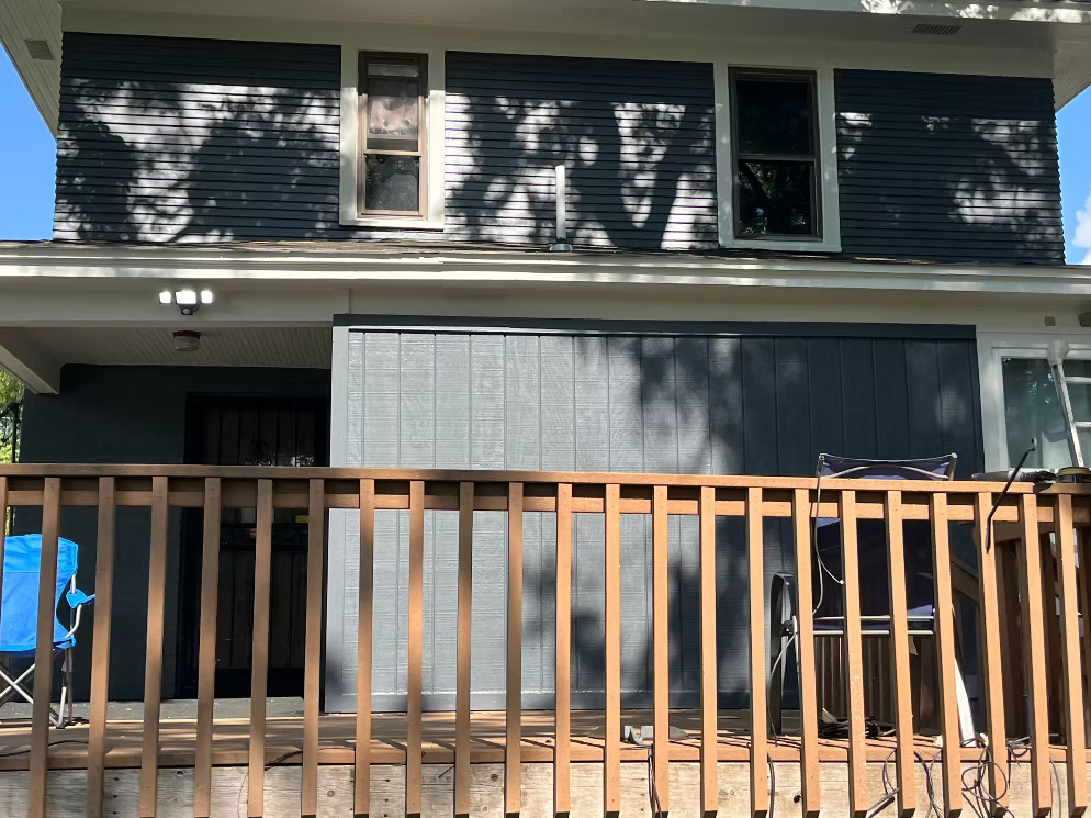 Front view of a house with blue siding, a porch with a wooden railing, and two chairs, one blue and one black, placed on the porch. There are shadows of trees on the house, and windows on the upper and lower levels.