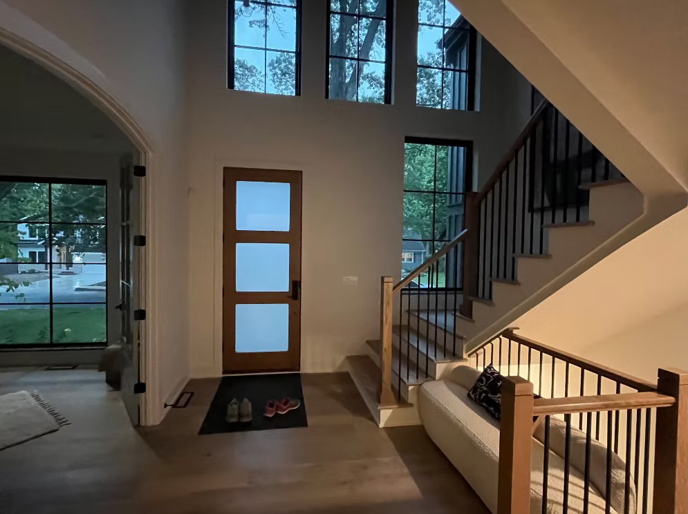 Entryway of a house with a wooden door, staircase with wooden railing, large windows, and a beige sofa.