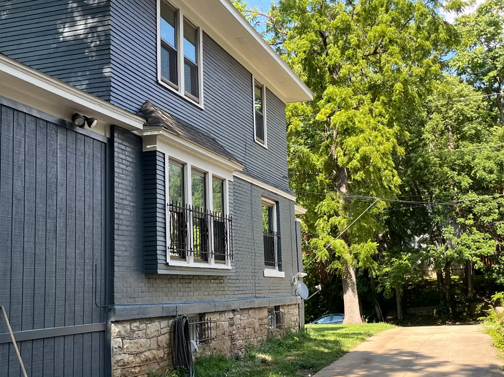 Side view of a modern multi-story house with blue siding, white trim, and stone foundation, surrounded by green trees and a driveway.