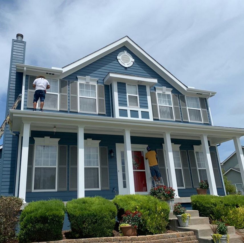 Two men painting the exterior of a large blue house with white trim, one on a ladder on the upper balcony and the other on the steps in front of the door, with a well-maintained garden and bushes in the foreground.