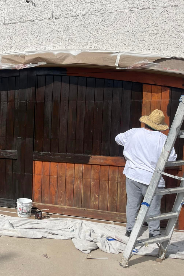 A person wearing a straw hat and white shirt painting a dark wooden fence, with a ladder leaning against it and painting supplies on the ground.