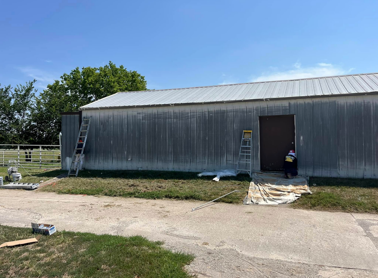 A worker in a hoodie applying paint to a metal building using a roller, with ladders on both sides, on a sunny day with a clear sky.