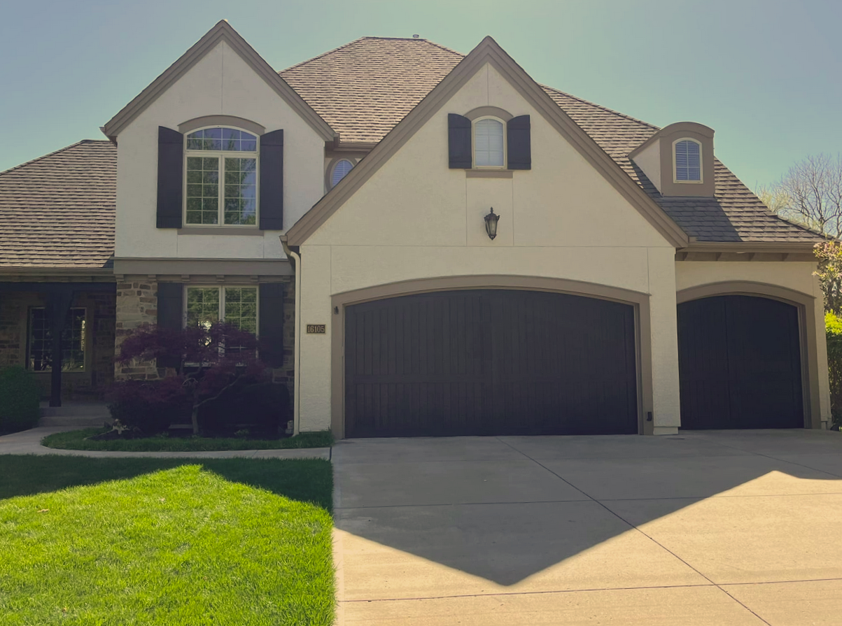 A two-story house with a beige stucco exterior, dark brown garage doors, black shutters, a stone accent wall on the left, a front yard with green grass, and a driveway.