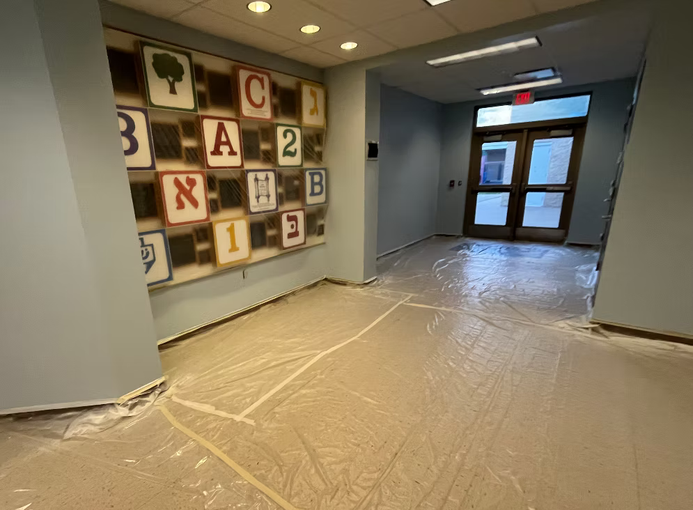 Interior of a building hallway with a colorful wall display of blocks with letters and numbers, plastic covering on the floor, and a door at the end of the hall.