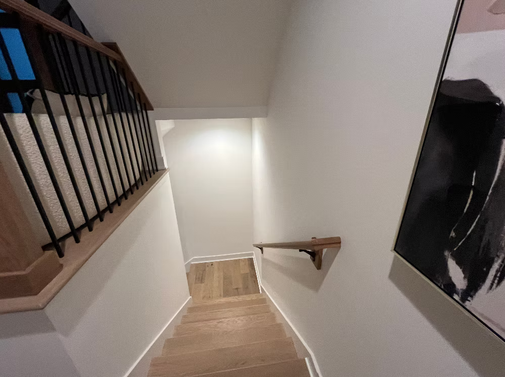 A staircase leading down to a small landing with wooden flooring, white walls, a wooden handrail, and a staircase railing on the left, with a large abstract framed artwork on the right wall.