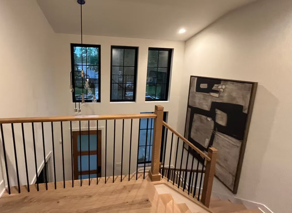 View from the top of a staircase in a modern home, showing three tall windows, a wooden railing, and a large abstract painting on the wall.